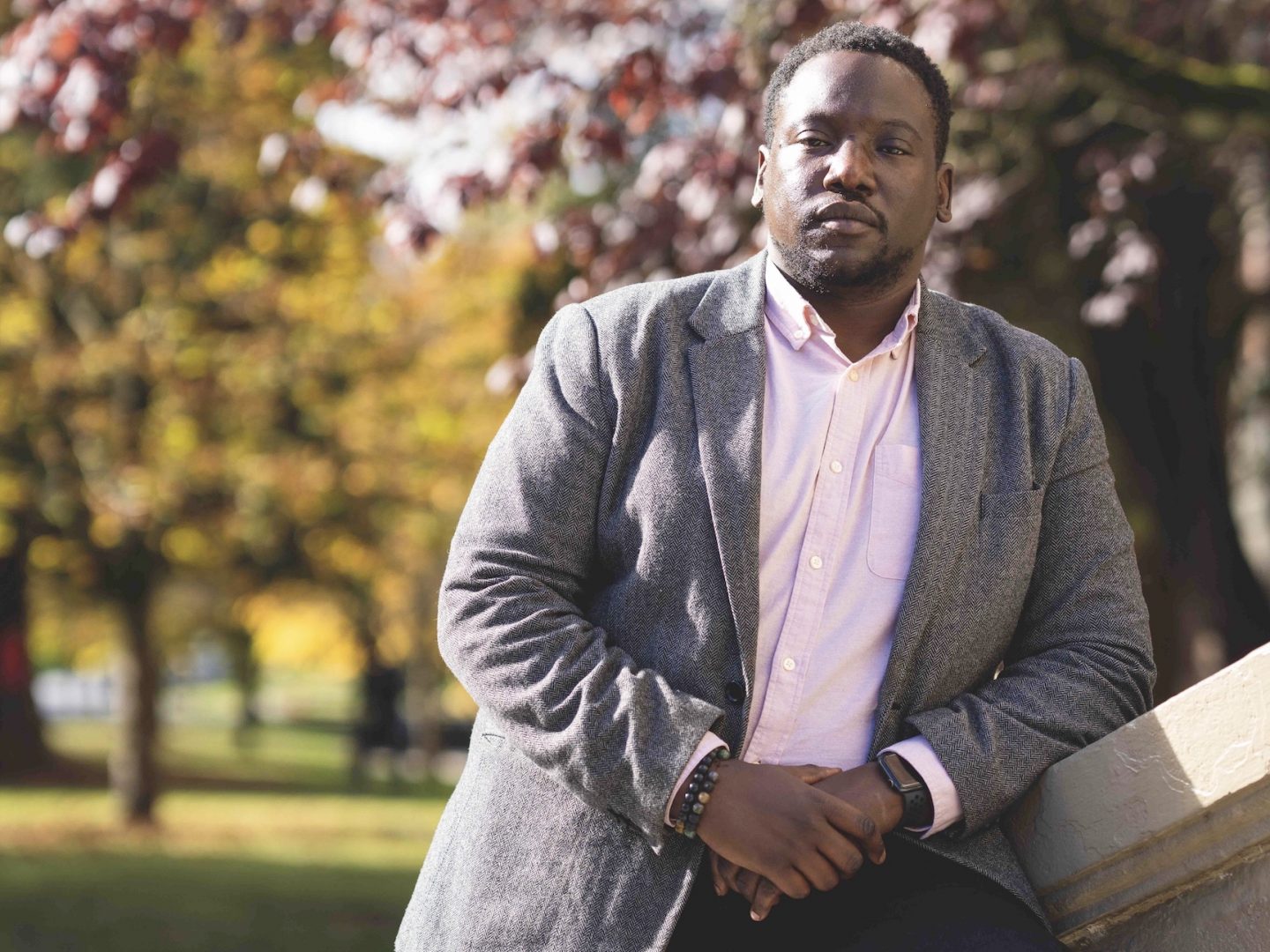 A Black man in a grey blazer leans against a railing.